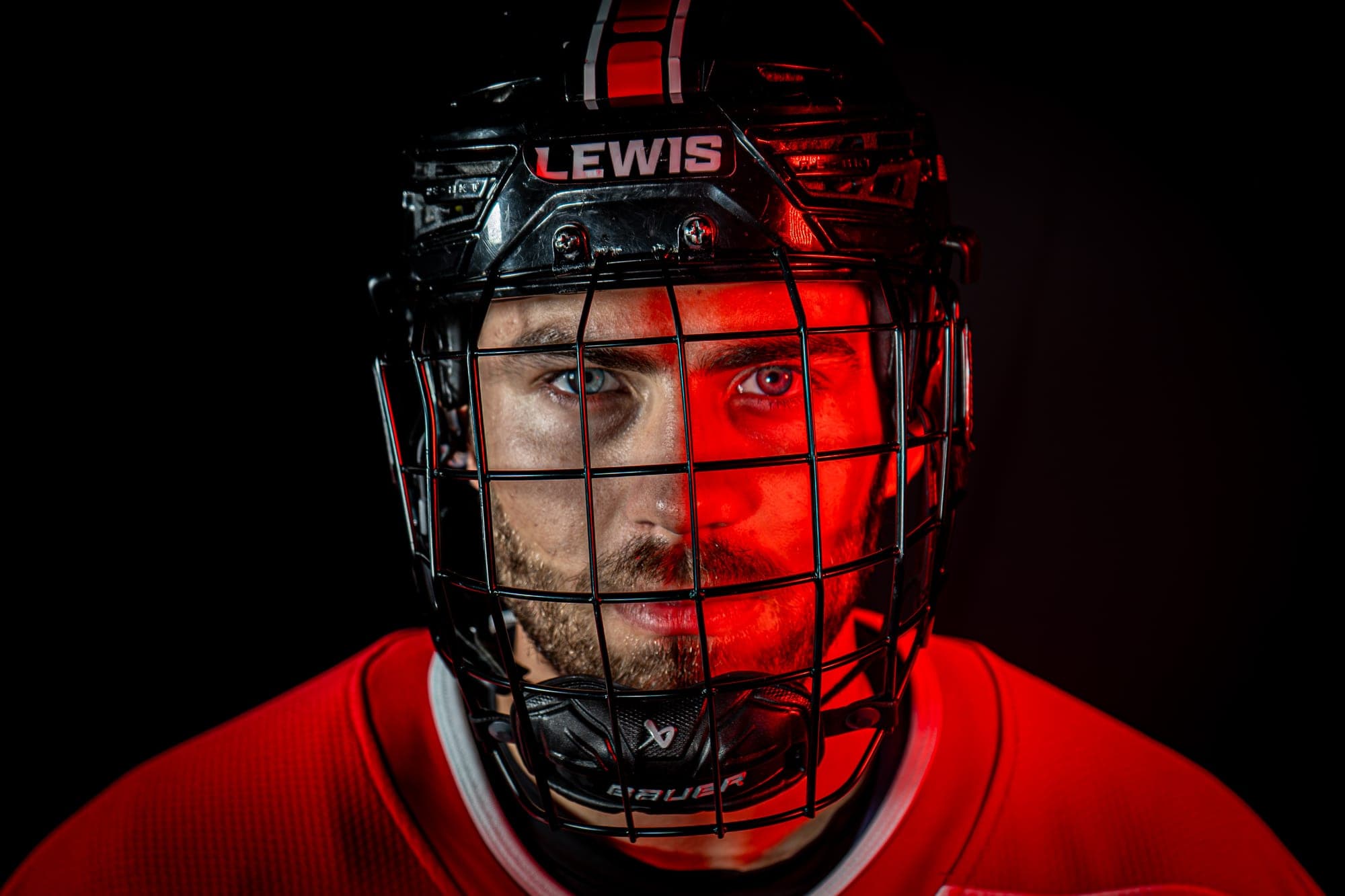 Close-up portrait of a Lewis hockey player in a caged helmet, half lit in red